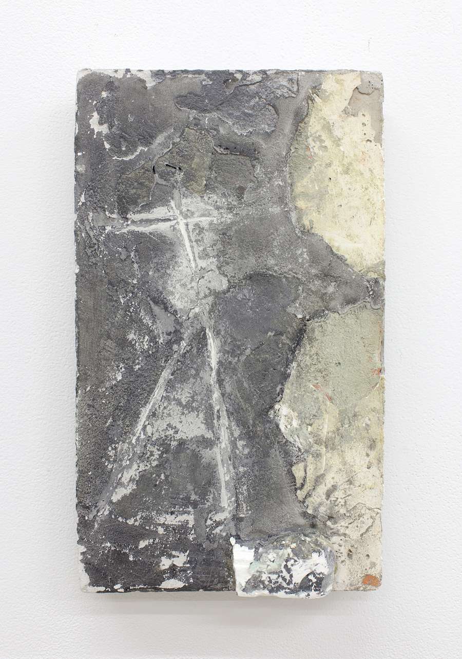 A cast concrete block is hung vertically on a white wall, the block is dirty and chipped with varying hues of gray and white. Carved into the surface is what appears to be the letters T and A stacked on top of each other.
