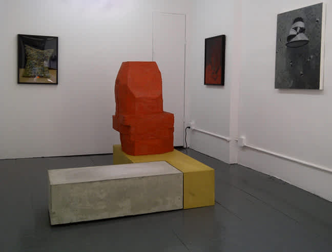 In a gallery space, three framed artworks are hung on white walls with a large floor sculpture resting on a wooden plinth in the middle of the room.