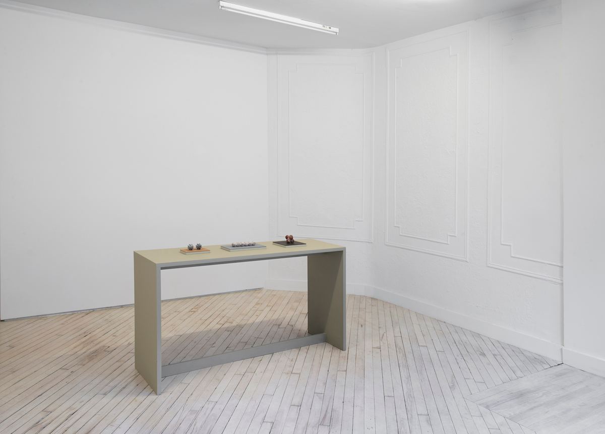 In a gallery space, a green tinted table stands in the center of the room. On top of the table are three plinths housing multiple, miniature sculptural objects resembling organic forms.