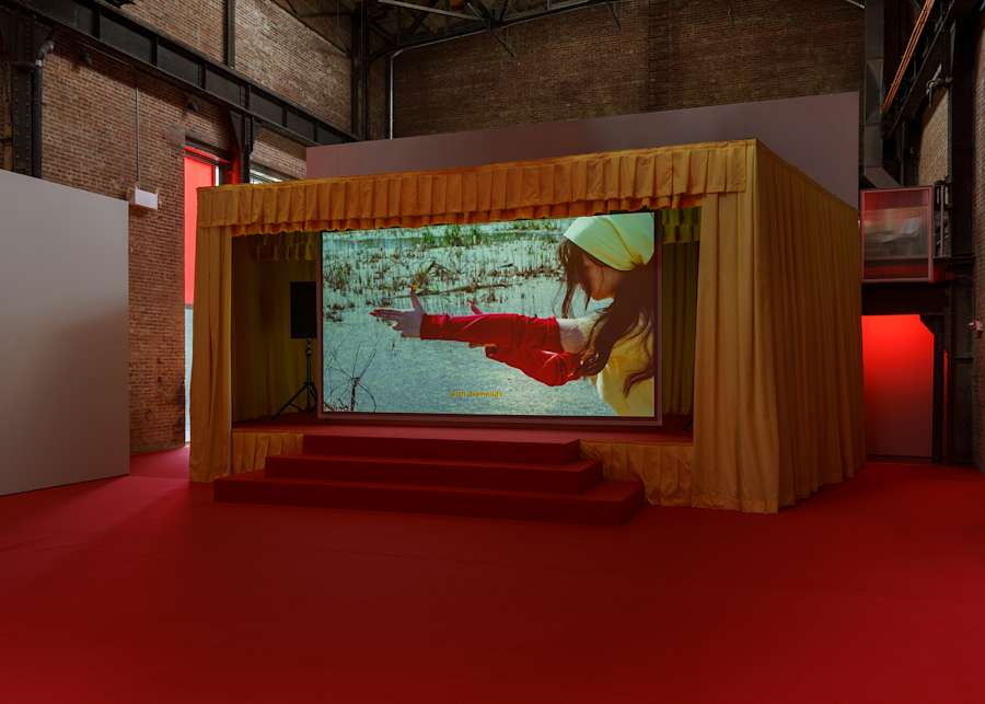A darkened brick gallery space with an installation resembling a red carpeted concert stage with yellow curtains. In the center of the stage is a large video projection of a young person holding out their arms in the shape of a gun with the words "with diamonds."