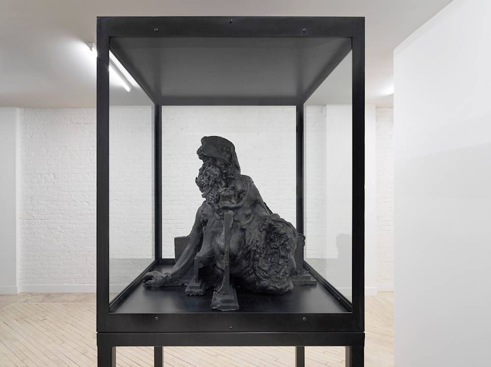 In a gallery space, a large metal and glass vitrine contains a cast iron sculpture resembling a decaying bust form.
