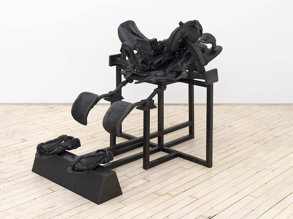 In a gallery space, a large cast iron metal sculpture rests on the floor. The sculpture resembles a riding saddle and accompanying straps on top of an architectural bracing structure.
