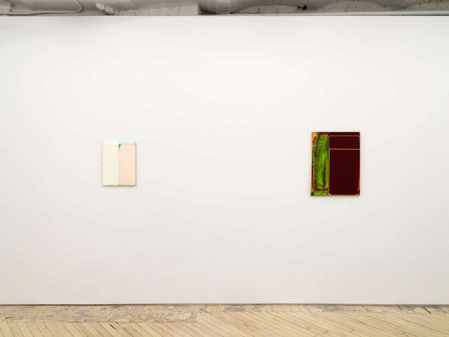 On a long white gallery wall, two abstract painting hung spaced apart. The paintings are abstract and feature blocked out colors in thick paint. The painting on the left is primarily off-white. The painting on the right is maroon and green.