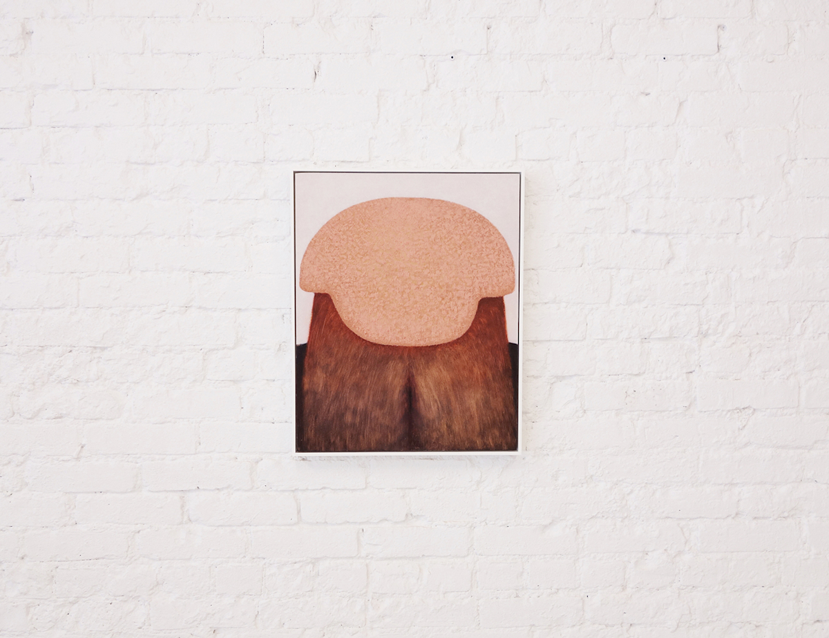 Installation view: Lui Shtini, Llapa, 2014: small painting in tones of peach and brown of a helmet-like head object that also looks like a bottom, framed in a thin white frame, hanging on white brick wall.