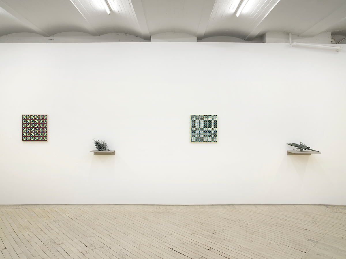 On a long white gallery wall, two square paintings depicting a grid of crisscrossing lines rendered primarily in blues, purples, and greens. separating the paintings are two abstract sculptures resembling archaic amalgamations of organic material. The sculptures are resting on top gray shelves attached to the wall lower than the paintings.