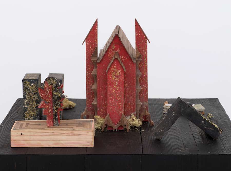 Detail close up of the top of a black pedestal showing red, black and wood objects, many of them covered lightly with a patina of yellow mold. At center is a model of a simple, red gothic church, to the left and right are small models of well known modernist sculptures, by Calder, and Tony Smith.