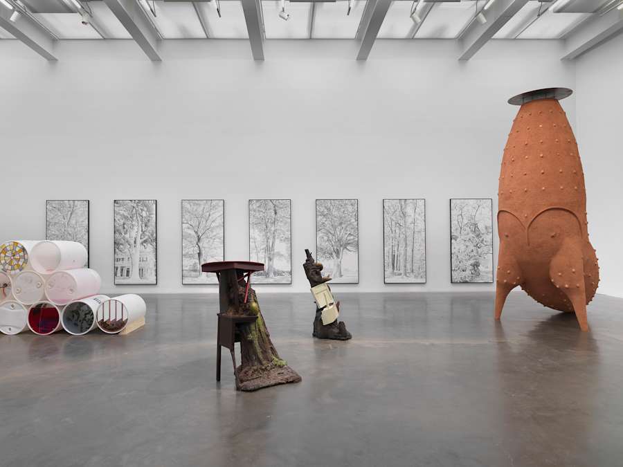 In a gallery space, a series of containers are stacked on top of each other to the left. In the background along a long wall are a series of large black and white paintings of tree. To the right are large freestanding sculpture nearly touches the ceiling. In the center of the room are two free standing sculptures resembling tree stumps combined with scraps of household furniture.