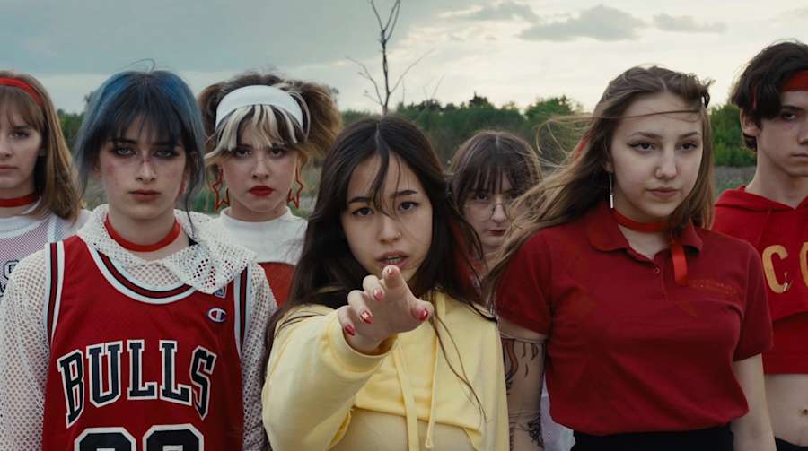 A close-up still image of a row of teenagers wearing matching outfits in reds and yellow. The center figure is pointing and looking directly into the camera.