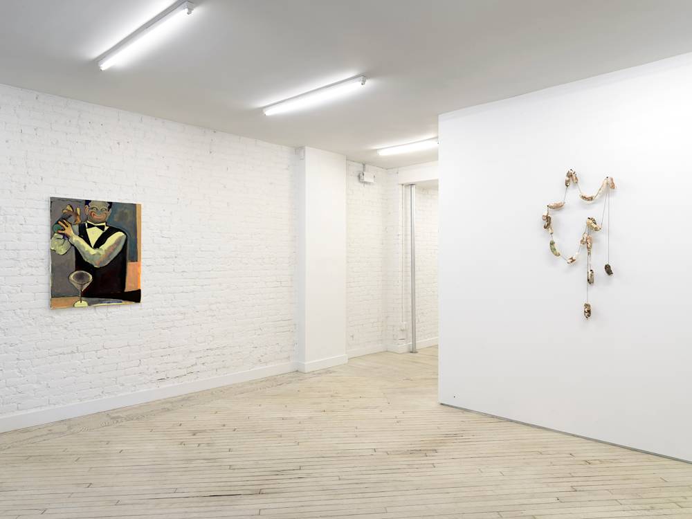 A gallery space with a row of neon lights hanging from the ceiling. On the left wall is an abstracted painting of a bartender shaking a drink with a clown-face expression. On the right wall is a wall hanging sculpture consisting of many sausage links arranged in an irregular order.