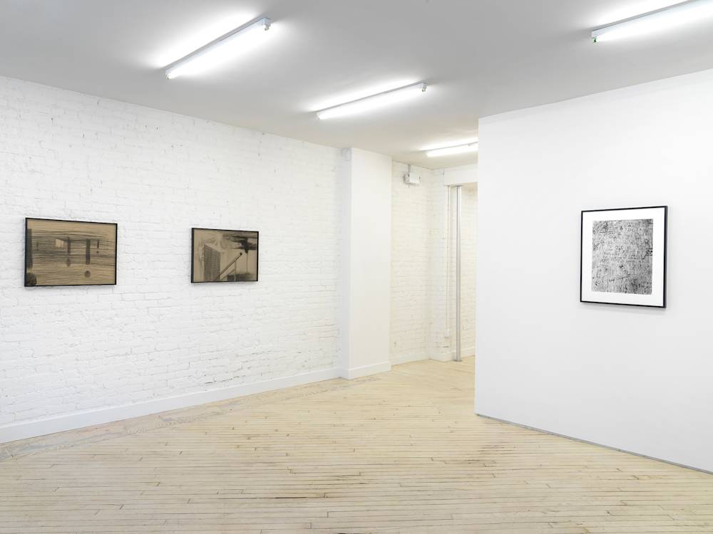 A gallery space with two pencil drawings behind tinted glass frames. To the right is a framed black and white photograph resembling a bird's-eye view of a wooden tabletop with numerous signs and marks carved into it.