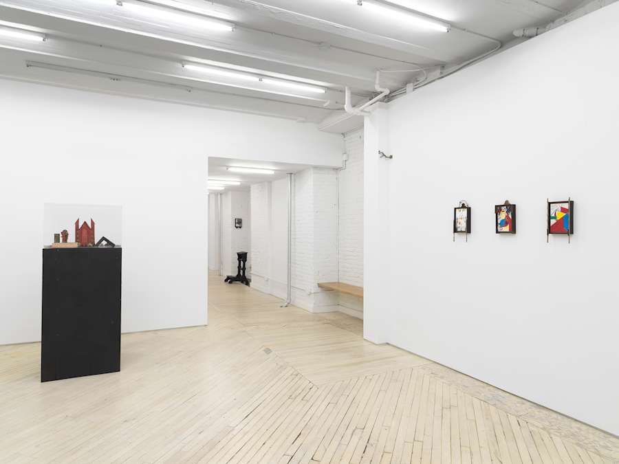 Installation view looking out from the center of the gallery towards the hallway. At left a black plinth with small red and black objects placed on top inside a glass vitrine. At left three small colorful framed drawings on the wall.