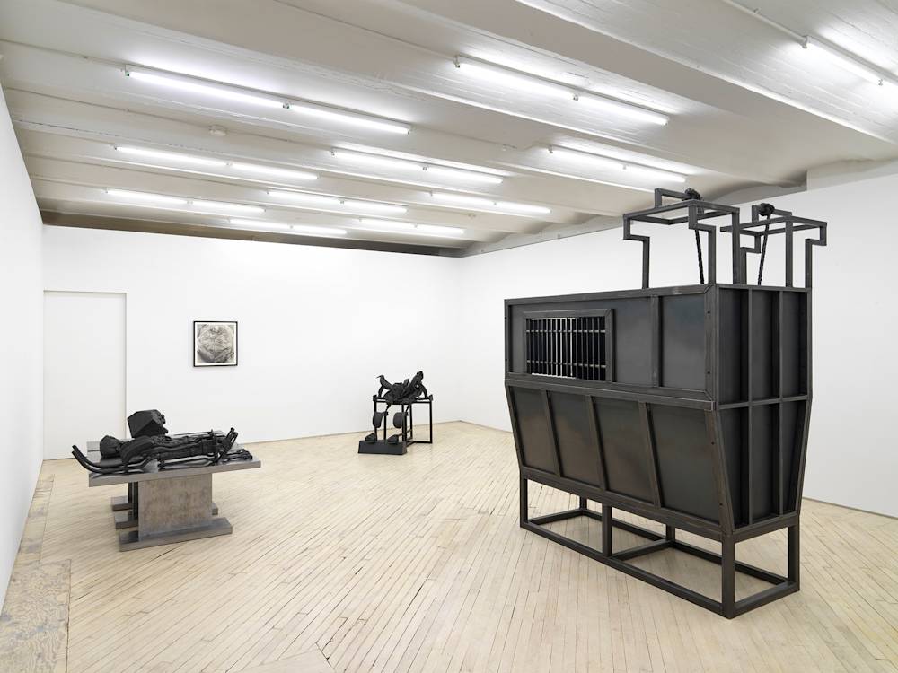 In a gallery space, a large metal and steel sculpture rests on the floor to the left. In the middle is a graphite drawing hung in black frame next to another iron sculpture. To the right of the gallery is a large steel structure resembling a cattle container.