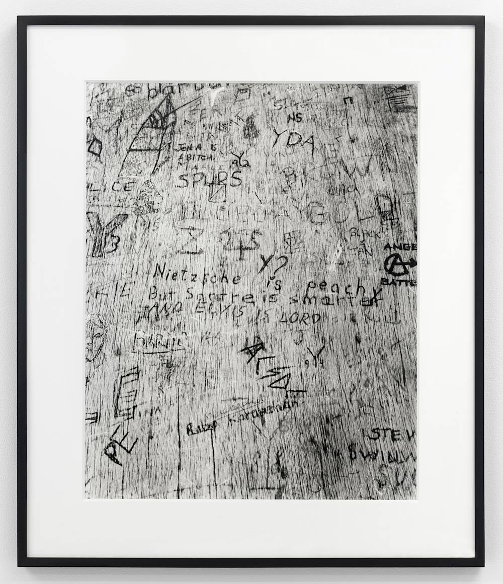A black and white photograph depicting a bird's-eye view of a wooden desktop with numerous, carvings or marks throughout the surface. The photograph is in a black frame.