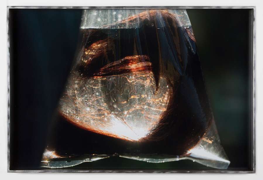 A clear plastic bag filled with water hangs in front of the camera, lit from behind, showing an eel like mass made of hair suspended in the bag.