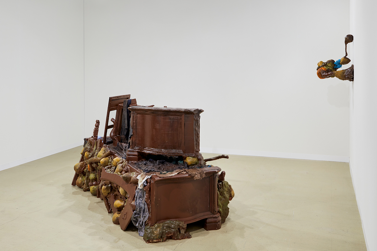 A large piece of brown furniture sits in the middle of a room with white walls. A smaller sculpture hangs to the right of the larger installation on the wall.