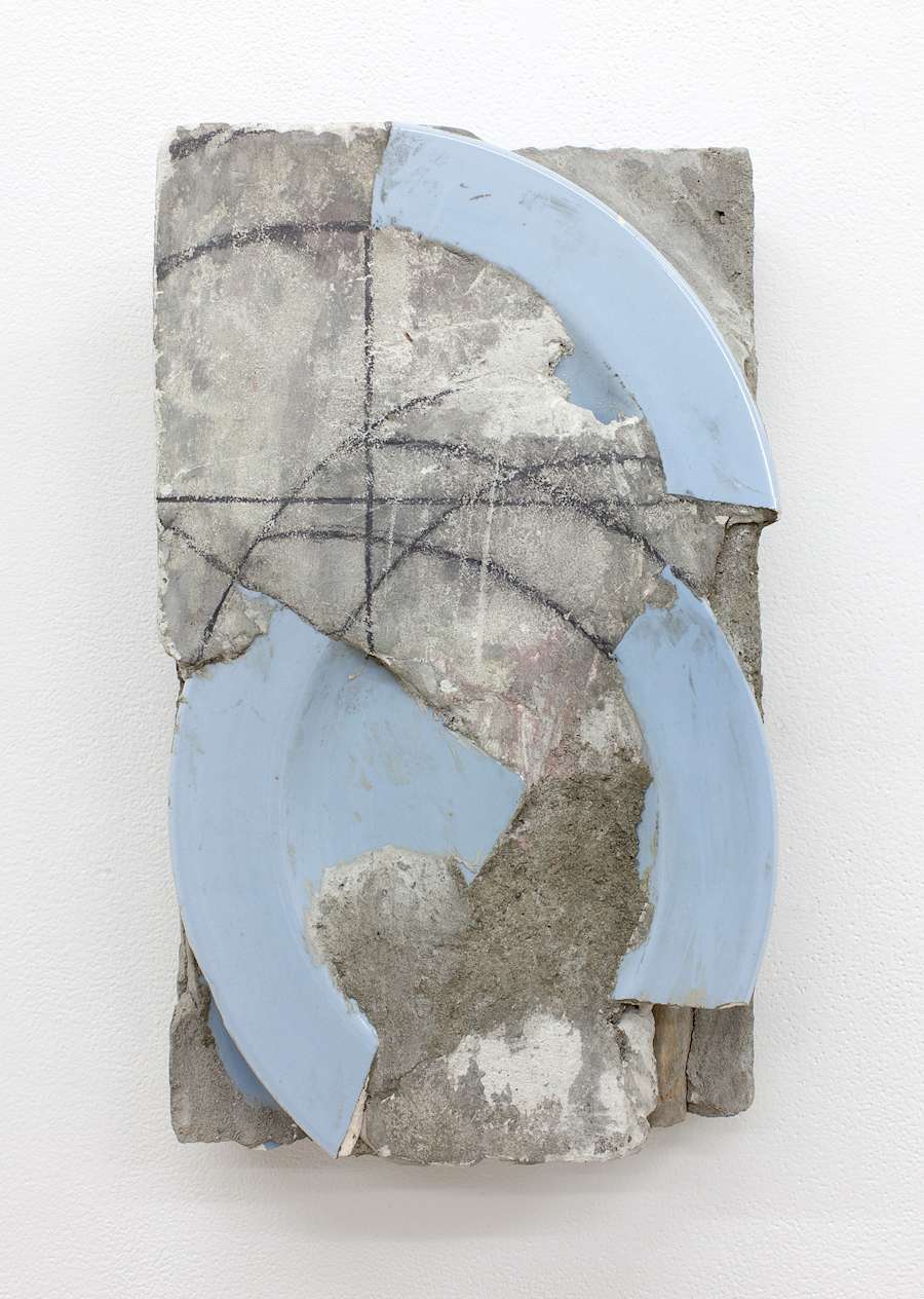 A cast concrete block is hung vertically on a white wall, the block is dirty and chipped with shards of a blue plate embedded in it. There are circular drawing marks on the surface of the concrete.