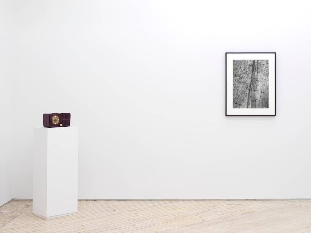 In a gallery space, a vintage purple radio to the left of a framed artwork resembling a black and white bird's-eye view of a wooden surface with numerous engravings or marks carved into the surface. The frame is black.