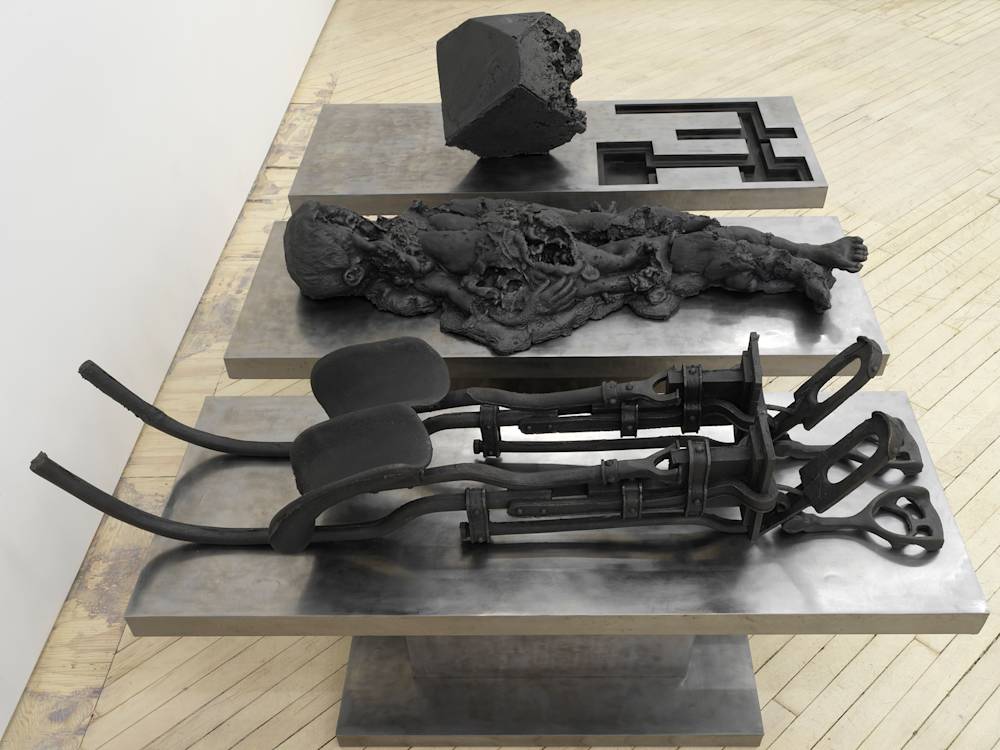 In a gallery space, a large cast iron metal sculpture rests on the floor. The sculpture is made up of multiple components. Cast iron forms resemble a contraption device, an abstracted body, and a cubic form.