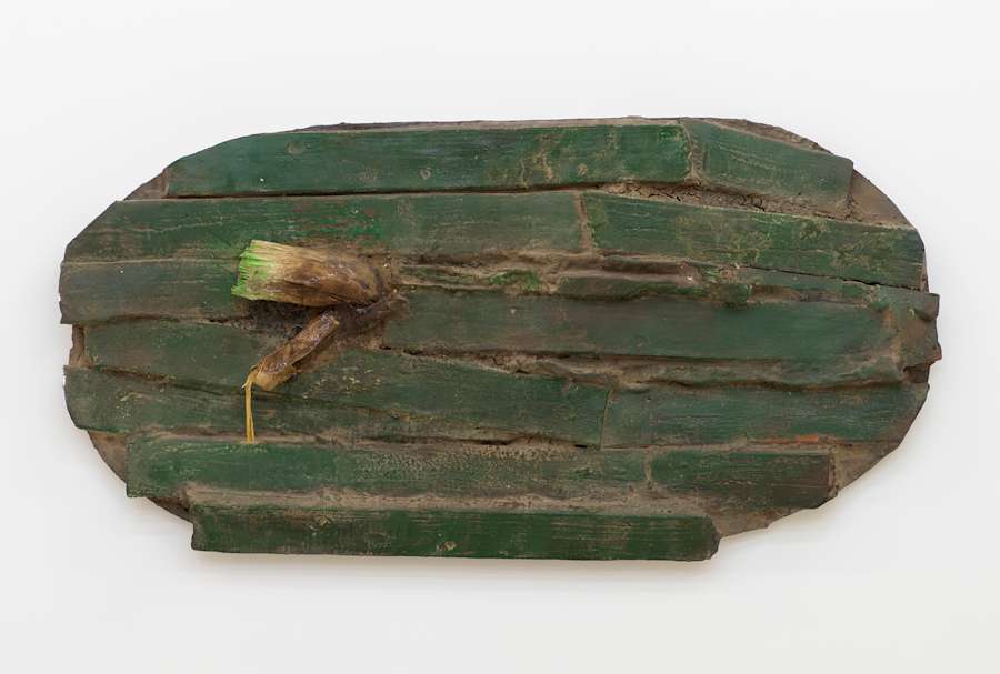 On a white wall, a rounded wall hanging sculpture consisting of numerous strips of irregular wood paneling painted green. To the left of the object is a cast vegetative-like protrusion.