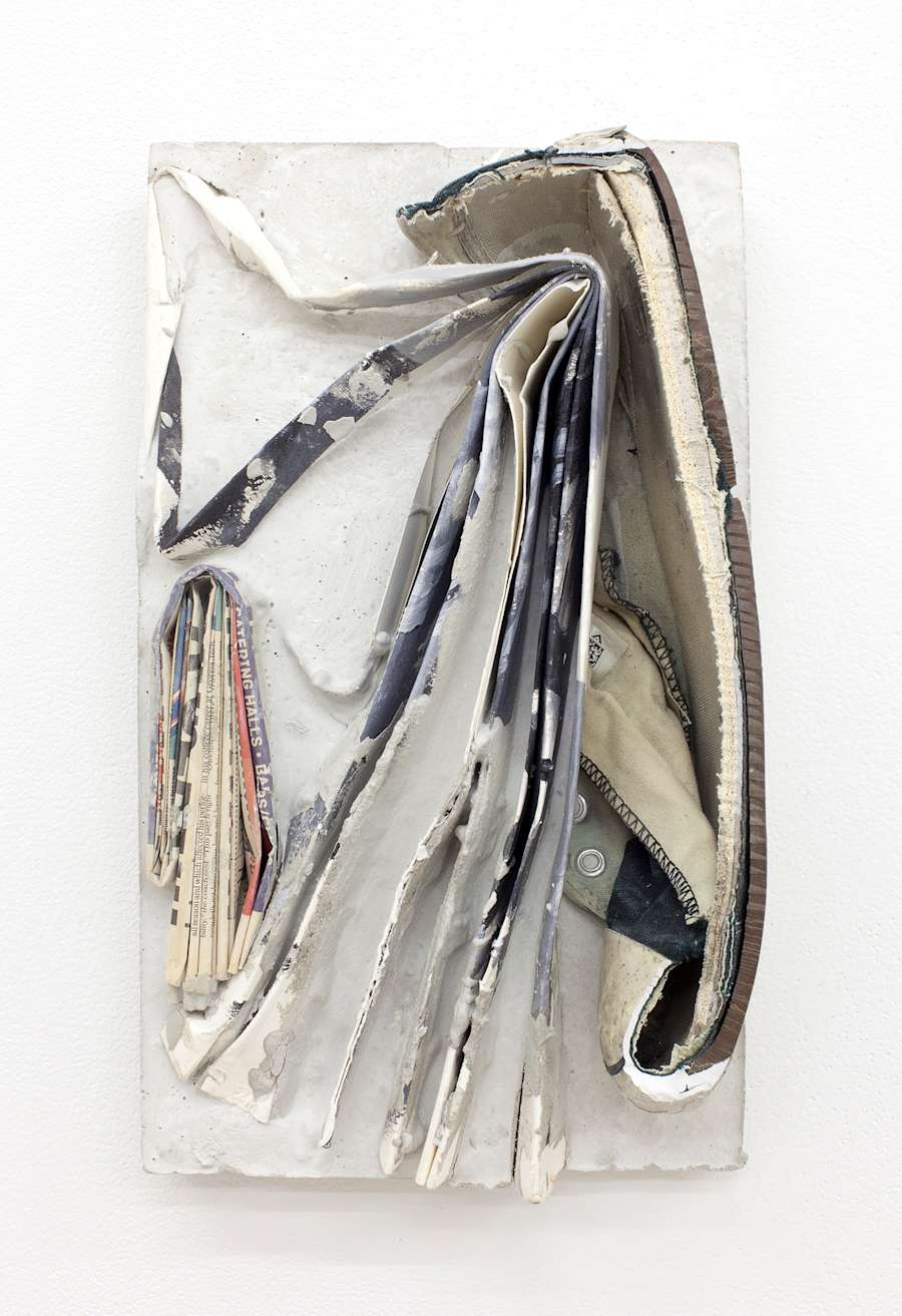 A cast concrete block is hung vertically on a white wall, the block is dirty and chipped with a Converse sneaker cut in half and embedded in the concrete, there is also a folded up newspaper embedded in it.