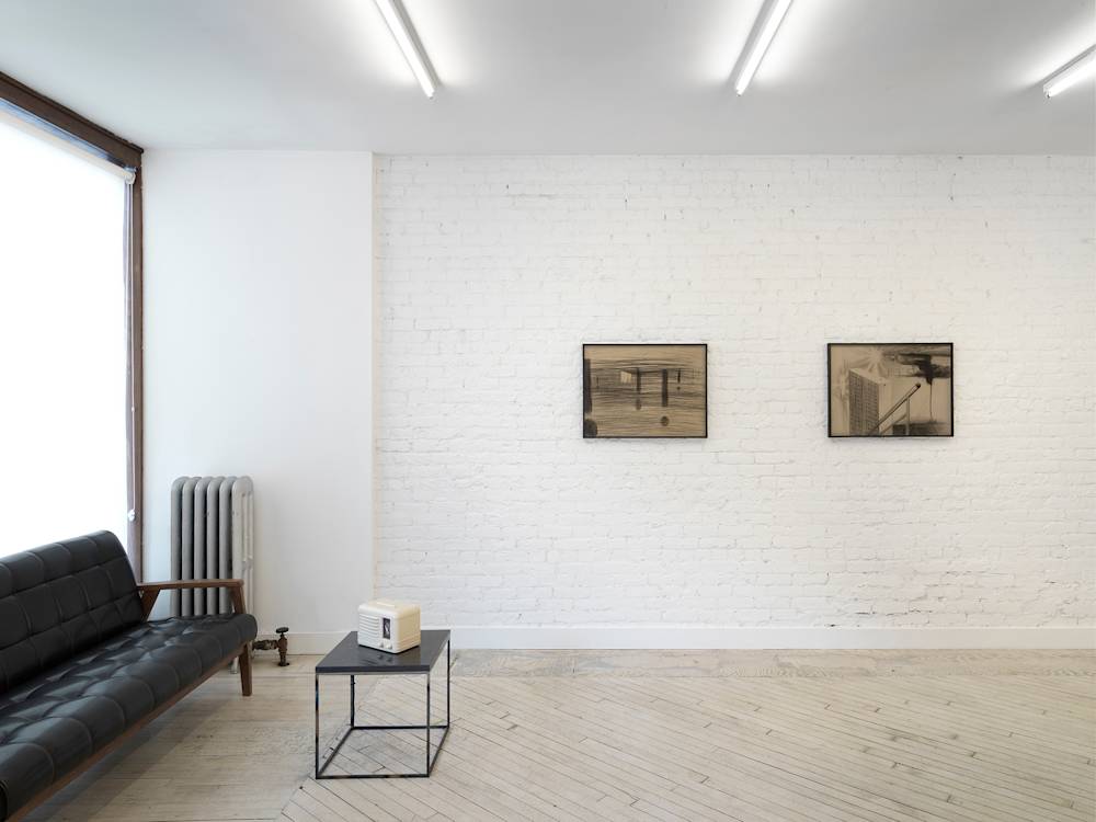 In a gallery space, a black leather couch with a small black table in front of it. On top of the table is a white vintage radio. On the center wall are two pencil drawings behind tinted glass frames.