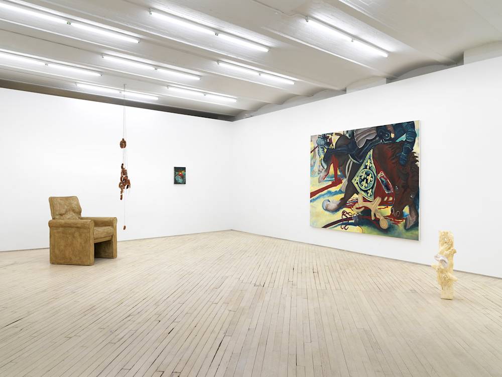 In a gallery space, a large brownish sculpture of a chair is placed next to a sculpture consisting of sausage links hanging from the ceiling. To the right is small, dark painting. On the right wall is a large abstract painting of a horse in the middle of a battle scene. On the floor to the right is an abstract sculpture with a light-brown finish. The sculpture has a sock covering one of the protrusions.