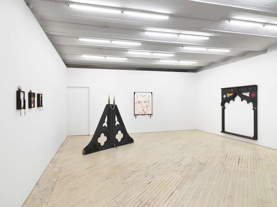 Installation view of main gallery: at left three small colorful drawings hang, framed with ornate black wood, a pyramid shaped gothic style wooden sculpture sits at center left behind which is a black and red drawing hanging on the center wall, framed as well in black wood, at right a large wooden archway form hangs on the wall.