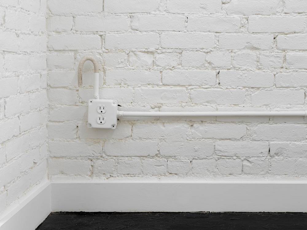 In a gallery space, a piece of conduit extending out of an electrical socket. In the background is a brick wall painted white.