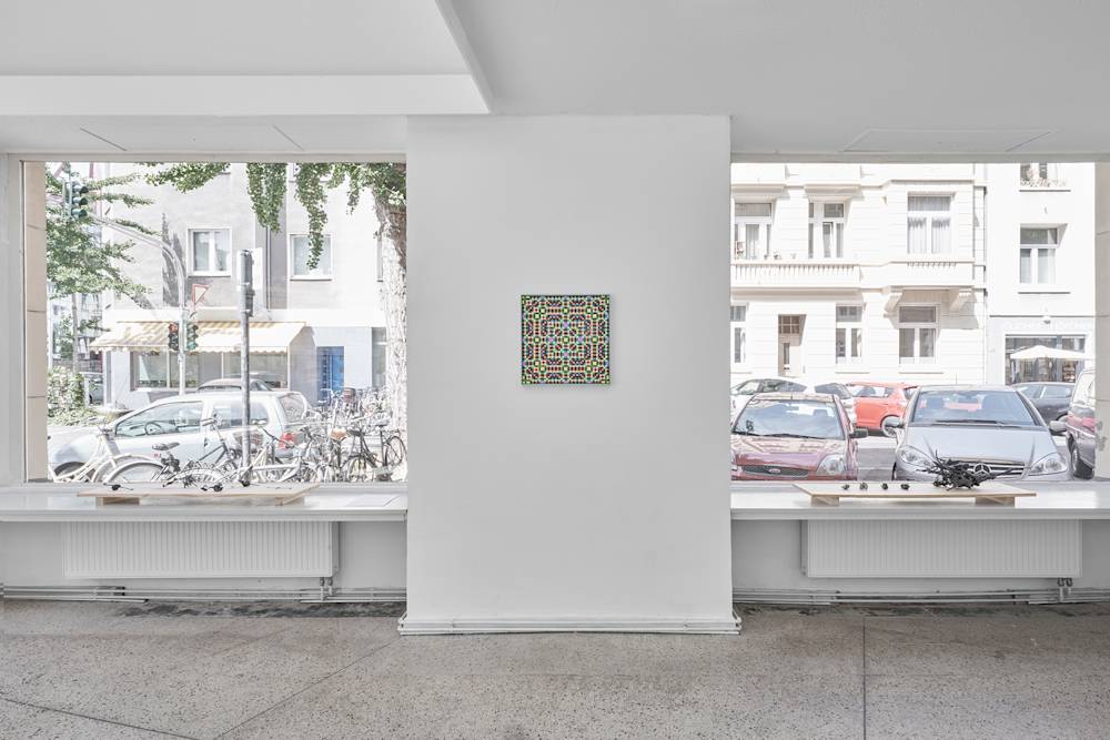 Image of a gallery room with two large windows, opening into a street view with parked cars and many bikes, with a wall in-between the large windows. A painting is installed on this wall. Two displays of the sculptures on a wooden slab are put on both sides of the wall, in front of the window.