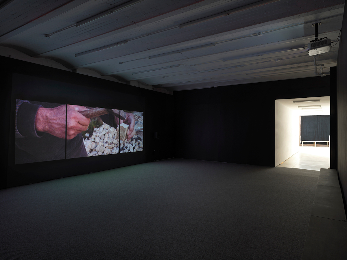 View of Gallery space with Video Installation projected onto dark wall, which exits into a lit hallway. Projected is a still of a hands shaping a piece of limestone with a small hammer.