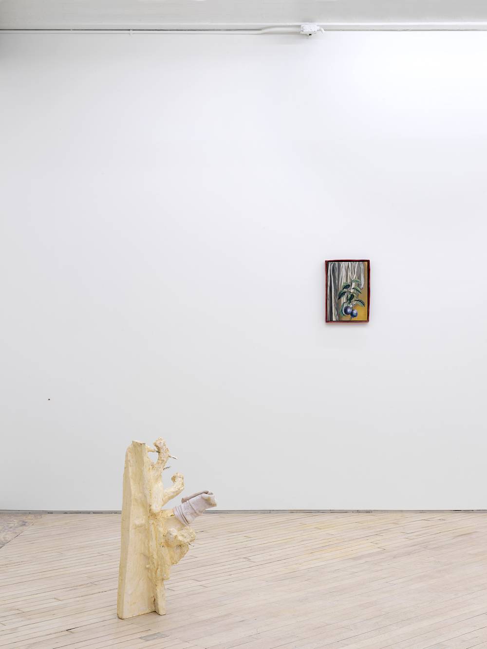 In a gallery space, an abstract irregularly shaped sculpture freestanding on the ground. The sculpture has a light-tan finish and multiple protrusions. One of the protrusions has a sock covering it. In the background is a painting depicting blueberries and a stem in an regularly shaped red frame.