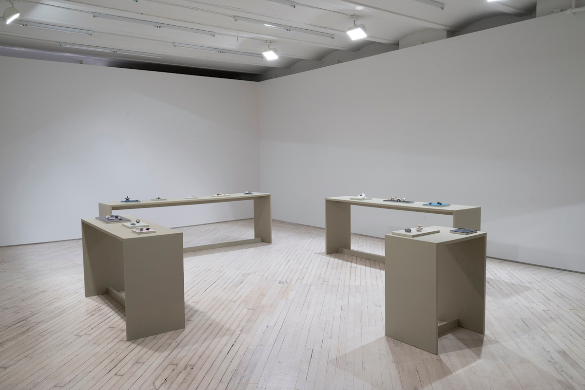 In a darkened gallery space, four tables arranged in a square facing one another. Resting on top of the tables are numerous small plinths housing various miniature sculptural objects.