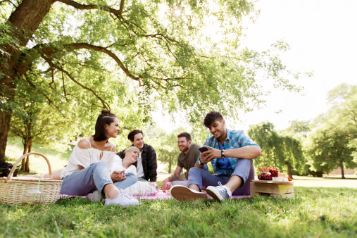 友情、レジャー、テクノロジー、人々のコンセプト – スマートフォンを持ち、夏の公園でピクニック用の毛布を冷やす友人のグループ