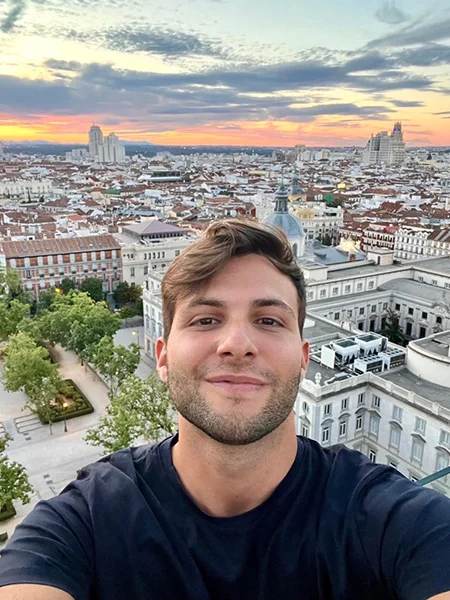 Una persona sonriente tomando un selfie con un horizonte urbano al atardecer de fondo