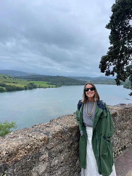 Mujer con abrigo verde de pie junto a un río, con colinas y cielos nublados