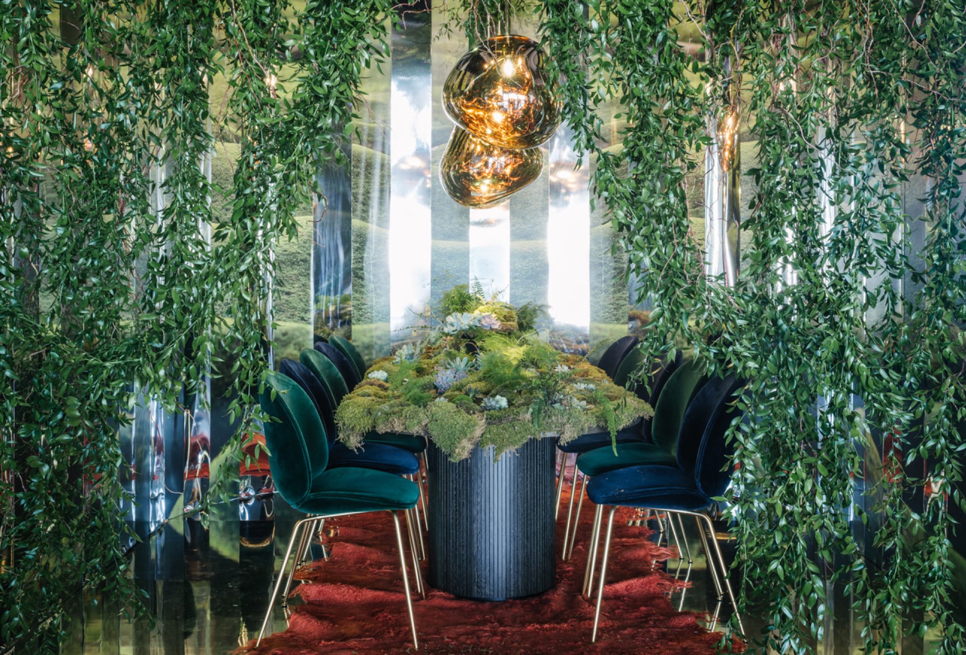 DWR for DIFFA lush, green fantastical room setting. Dining table covered in moss and ferns surrounded by velvet chairs. Greenery hanging from walls with strips of mirror. Golden amorphic hanging lamps over table.