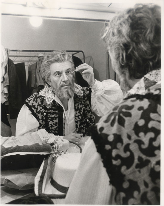 black and white photograph of a man wearing theatrical costume looking in a mirror while applying make-up