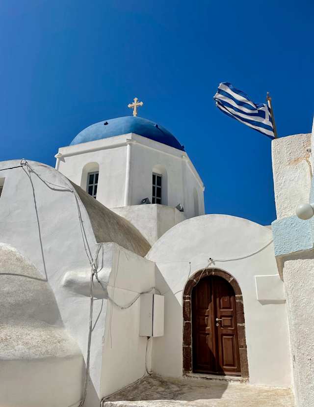 A blue-domed church with a Greek flag in Pyrgos Kallistis, Santorini, Greece