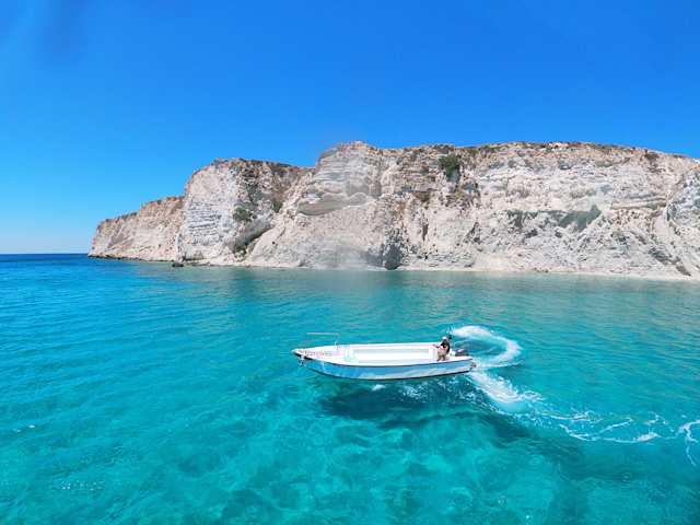 A white boat on clear blue water in front of a large rock in Crete, Greece
