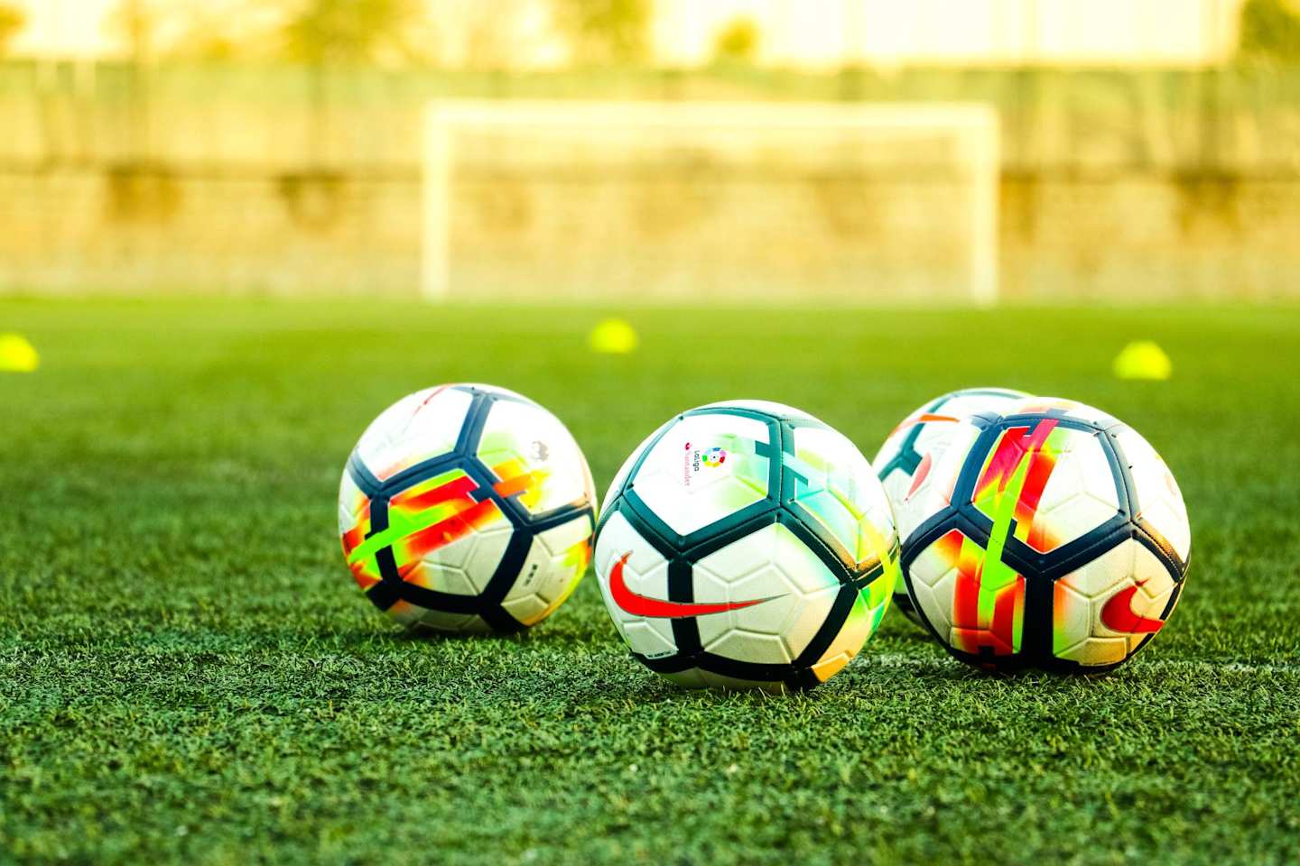 Four footballs on a football pitch in front of cones and a goal