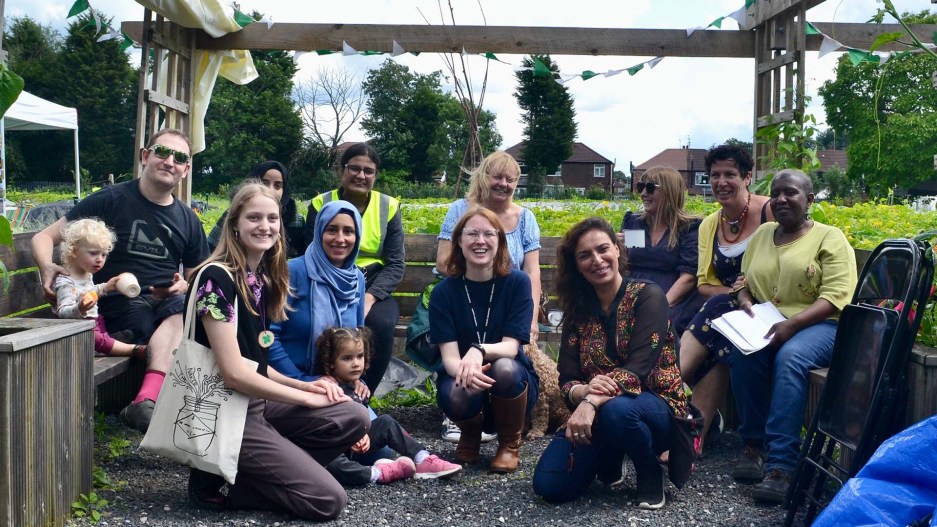 A group of people sat on an outdoor bench, smiling at the camera. There's a meadow and trees in the background.