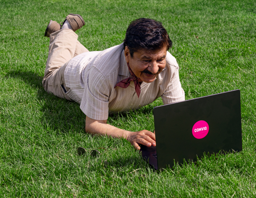 En leende äldre man ligger på grönt gräs och använder en svart laptop med en rosa Comviq-klistermärke.