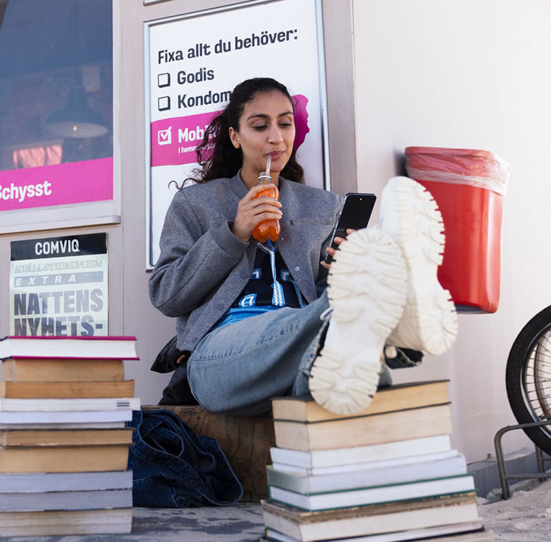 Nadja sitter utanför Karims kiosk med fötterna på en trave böcker, dricker något med sugrör och tittar på sin mobil