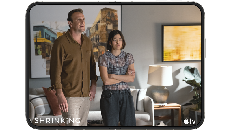 På en iPhone Duo-skärm ser man en man och en kvinna som står i ett vardagsrum i TV-serien Shrinking på Apple TV. Kvinnan har armarna i kors.