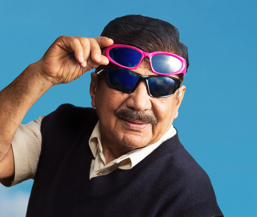 En äldre man med mustasch bär två par solglasögon utomhus mot en blå himmel med moln.