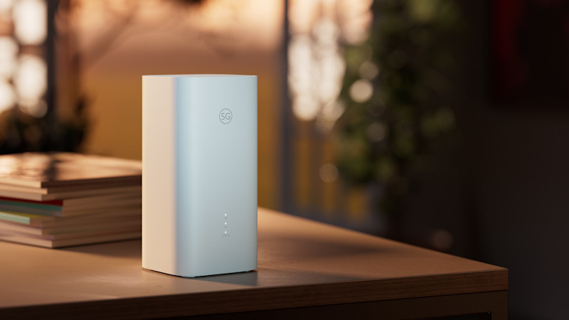 Vit 5G-router står på ett träbord i ett hemmiljö med mjukt kvällsljus i bakgrunden. Fyra små LED-lampor lyser på framsidan av enheten, och några böcker ligger bredvid på bordet.