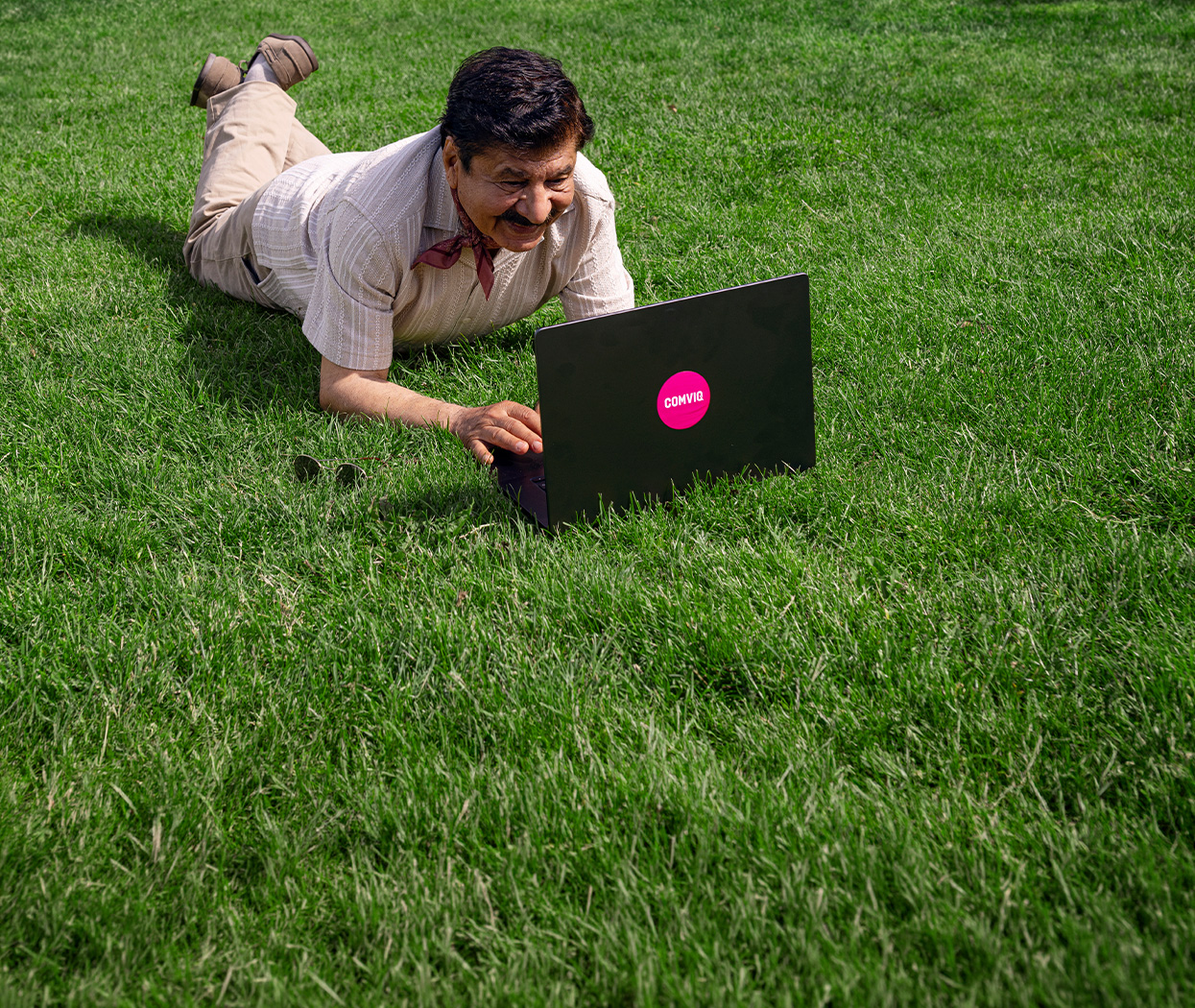 En leende äldre man ligger på grönt gräs och använder en svart laptop med en rosa Comviq-logga.