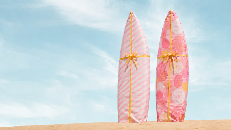 Två surfbrädor inslagna i rosa presentpapper. De står upprätt i sand med en blå himmel i bakgrunden.