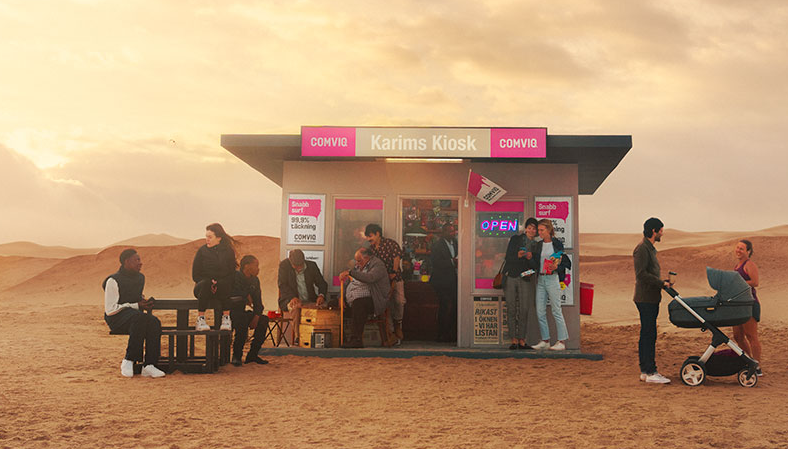 Bild på en kiosk det står "Karims kiosk Comviq" på placerad i en öken. Till vänster står ett bord där två män och en kvinna sitter, vid ingången till kiosken står flera människor. Till höger står en man och en kvinna med en barnvagn.