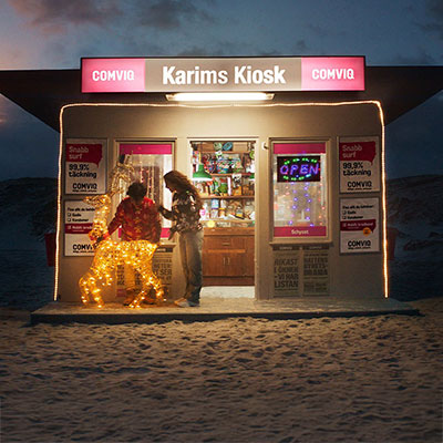 Upplyst kiosk i skymningen med Comviq-reklam och två personer vid ingången omgivna av ljusslingor.